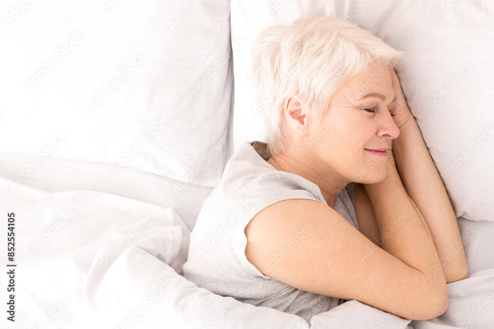 Fototapeta premium A woman is lying down in bed, covered by crisp white sheets. She appears relaxed and comfortable, with her eyes closed, possibly resting or sleeping, view above