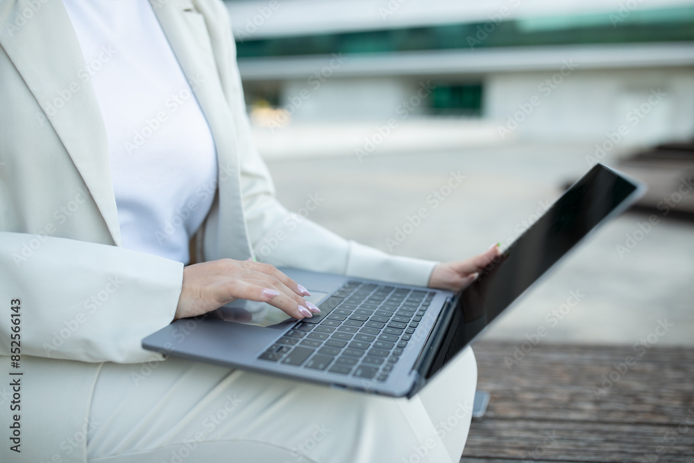 A woman wearing a white suit is sitting on a bench outside. She is using a laptop and appears to be working or browsing the internet. The background is blurred, suggesting a modern urban environment.
