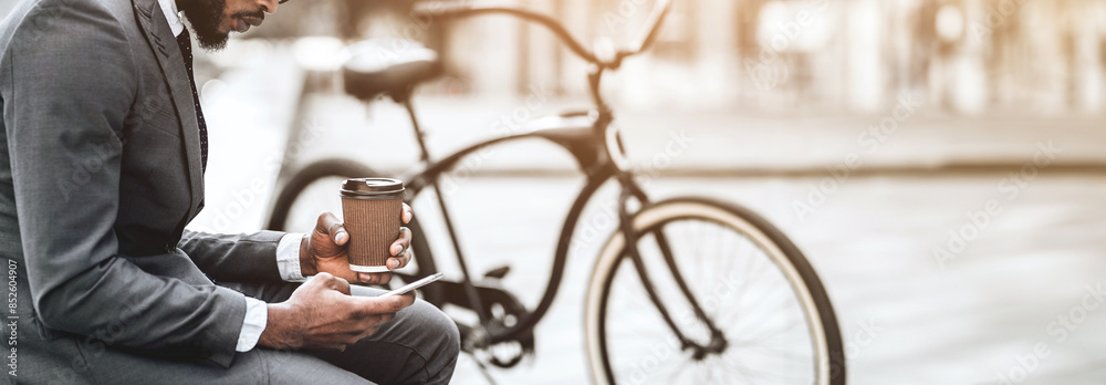Fototapeta premium Cropped of African American businessman holding coffee cup and phone, sitting near a bicycle in urban setting, panorama with copy space