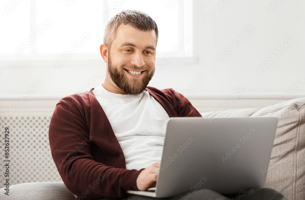 A man is seated on a comfortable couch, focused on a laptop screen in front of him. He appears engaged, possibly working or browsing. The room is well-lit, with a casual and relaxed atmosphere. A man is seated on a comfortable couch, focused on a laptop screen in front of him. He appears engaged, possibly working or browsing. The room is well-lit, with a casual and relaxed atmosphere.