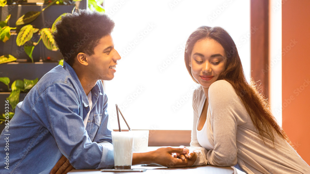 A young African American couple is sharing a moment of affection while enjoying smoothies at a cozy cafe, seated at a table, smiling and holding hands, with sunlight streaming through the window. A young African American couple is sharing a moment of affection while enjoying smoothies at a cozy cafe, seated at a table, smiling and holding hands, with sunlight streaming through the window.