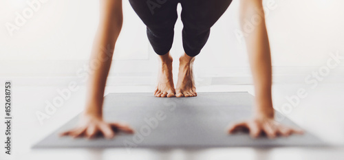 Unrecognizable woman is seen in a plank position on a yoga mat indoors. The image focuses on the legs and hands, suggesting a yoga exercise in a well-lit room during the day.