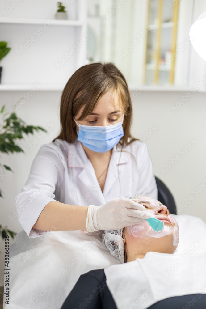 Portrait of a young cosmetologist wearing white gloves and a surgical mask. which is applying a clay mask to her client's face. Portrait of a young cosmetologist wearing white gloves and a surgical mask. which is applying a clay mask to her client's face.