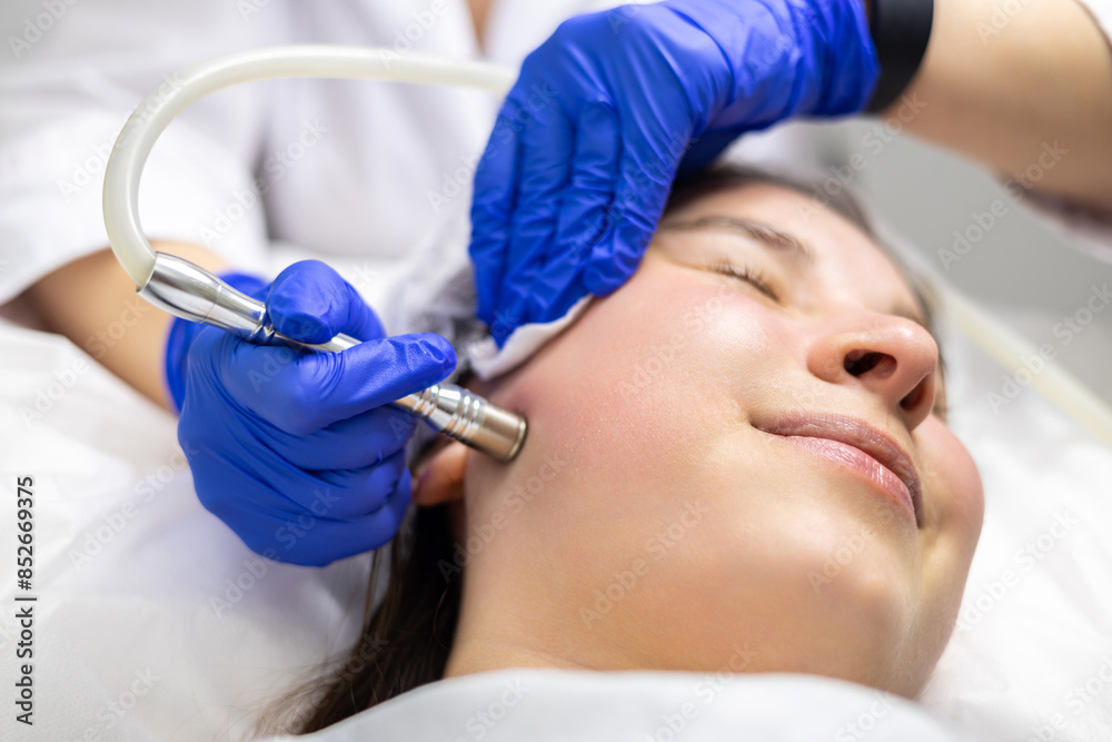 A professional dermatologist wearing medical gloves performs a microdermabrasion procedure on a smiling young woman in a professional beauty salon. A professional dermatologist wearing medical gloves performs a microdermabrasion procedure on a smiling young woman in a professional beauty salon.