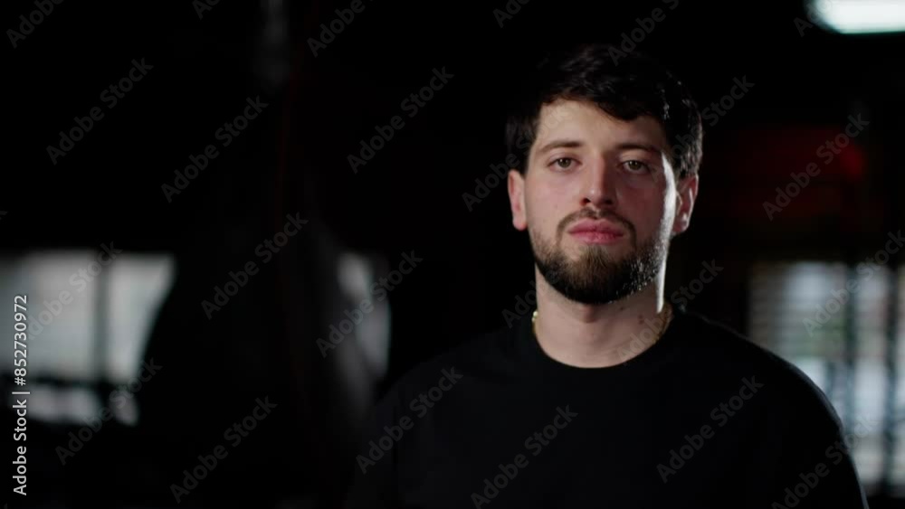 Young man with a beard and short dark hair standing in a dimly lit gym, wearing a black t-shirt, looking serious and focused. Young man with a beard and short dark hair standing in a dimly lit gym, wearing a black t-shirt, looking serious and focused.