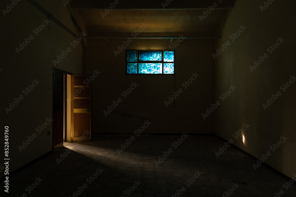 panoramica su una stanza vuota e buia di un edificio abbandonato, illuminata leggermente di lato dal sole da una porta aperta e di fronte da una finestra chiusa e di colore blu