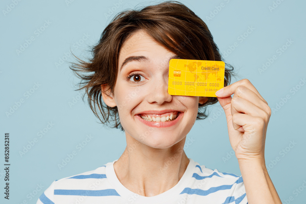 Close up young smiling woman wears striped t-shirt casual clothes hold in hand cove eye with mock up of credit bank card isolated on plain pastel light blue cyan background studio. Lifestyle concept.