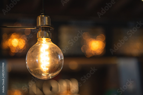 Fényképezés A classic designed tungsten lightbulb is glowing in orange warmlight shade with blurred background of building interior