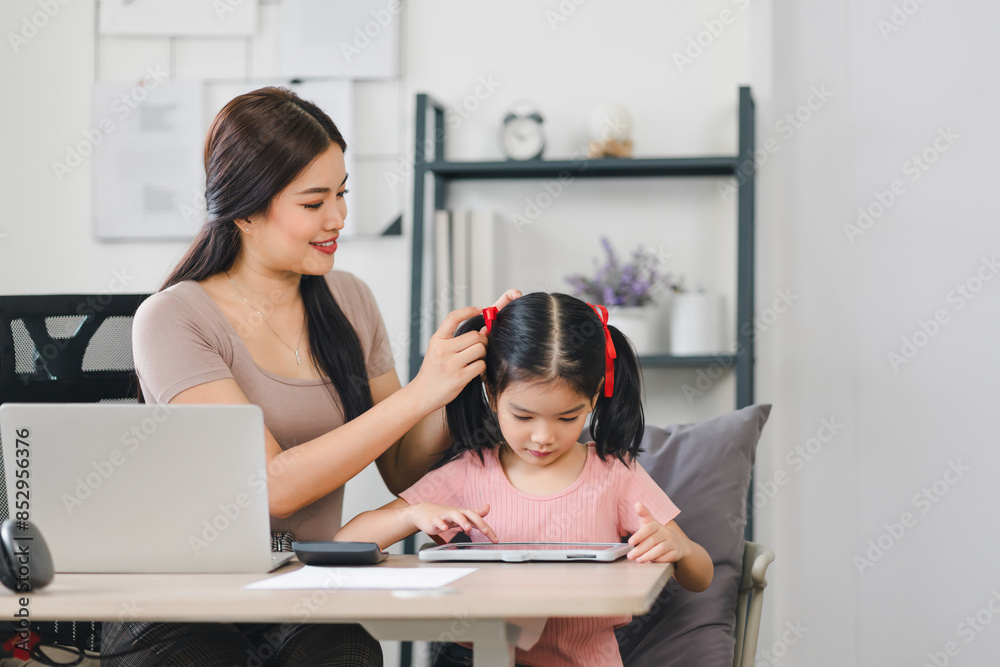 Mother working from home while helping her daughter with a digital tablet, balancing work and family time in a modern home office. Mother working from home while helping her daughter with a digital tablet, balancing work and family time in a modern home office.