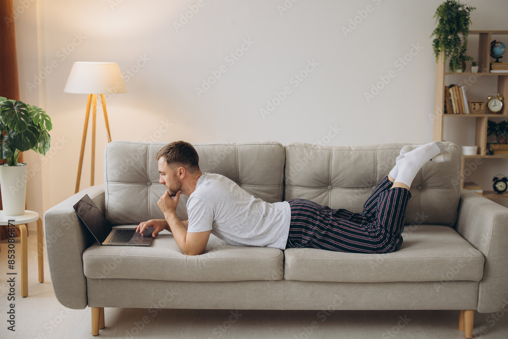 One man Caucasian male freelancer working from home with headphones on laptop computer customer service manager lying on sofa at home wearing shirt and pajamas One man Caucasian male freelancer working from home with headphones on laptop computer customer service manager lying on sofa at home wearing shirt and pajamas