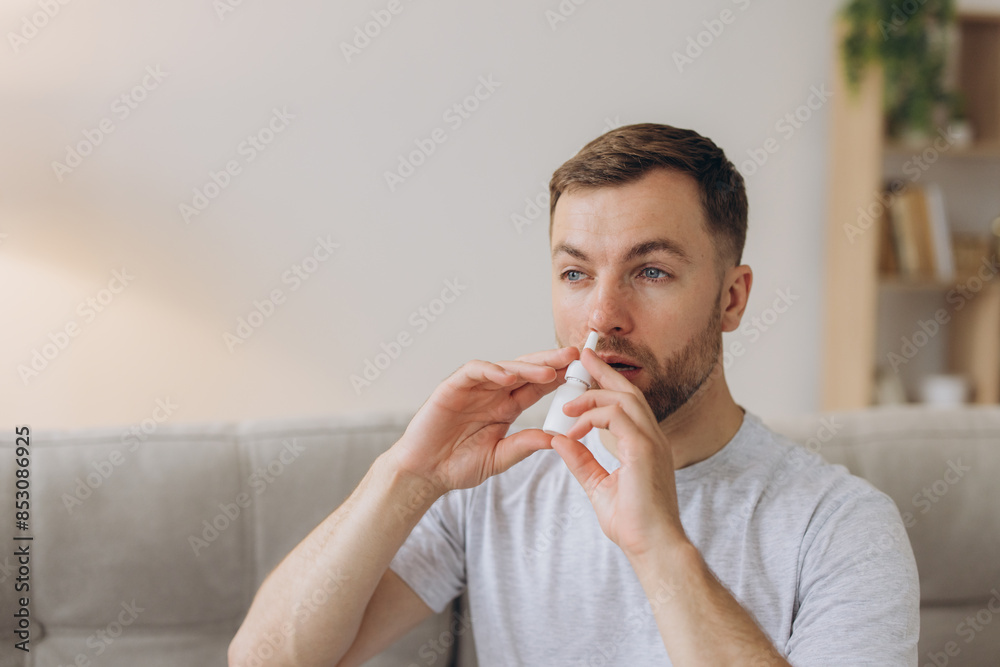Frustrated sick unhealthy ill allergic man has red eyes runny stuffy sore nose suffer from allergy symptoms hay fever uses nasal drops while sitting on sofa at living room Frustrated sick unhealthy ill allergic man has red eyes runny stuffy sore nose suffer from allergy symptoms hay fever uses nasal drops while sitting on sofa at living room