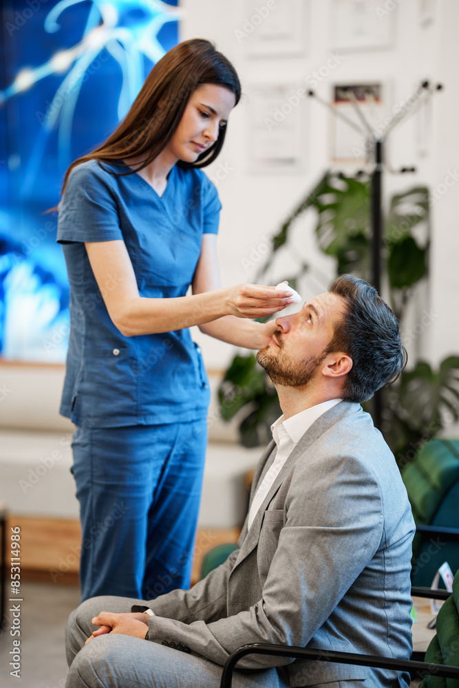Fototapeta premium Beautiful medical technician applying pupil dilation drops to a handsome male patient's eye before an eye exam. Modern clinic for diagnosis and treatment of eye and sight diseases.