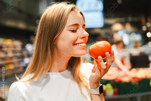 Smiling young woman choosing perfect tomato in grocery store