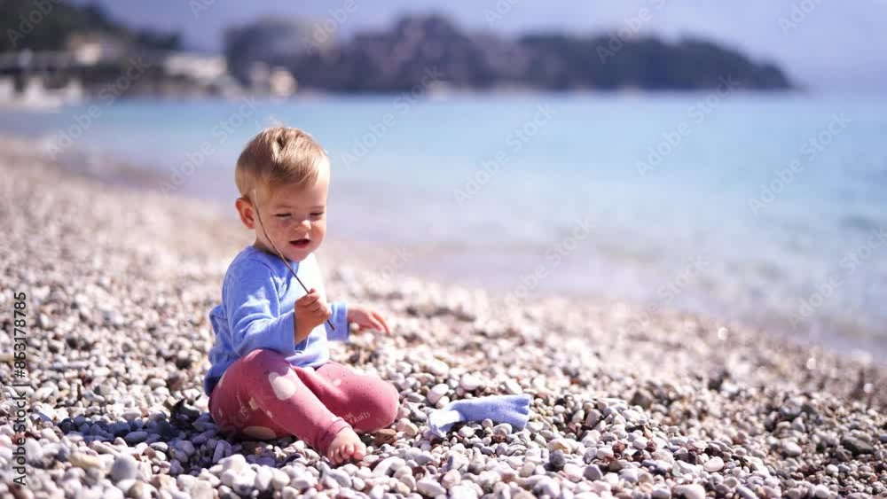 Little baby playing with a stick sitting on a pebble beach by the sea. High quality 4k footage Little baby playing with a stick sitting on a pebble beach by the sea. High quality 4k footage