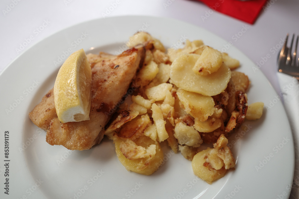 A delicious plate featuring a piece of pan-fried fish served with a lemon wedge and a side of sautéed potatoes. A delicious plate featuring a piece of pan-fried fish served with a lemon wedge and a side of sautéed potatoes.