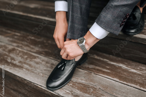 Stylish fashionable male businessman, young groom in a gray suit fastens black leather shiny shoes with his hands while sitting on a wooden stair step. Close-up photography, lifestyle.