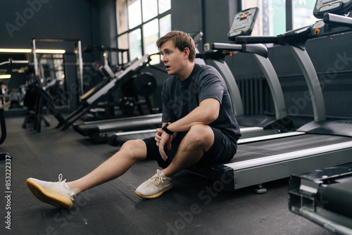 Fototapeta Naklejka Na Ścianę i Meble -  Portrait of tired runner taking break at sport gym sit on treadmill. Portrait of exhausted sweaty male hard breathing after cardio exercise running workout. Weary man sit on fitness treadmill relax.