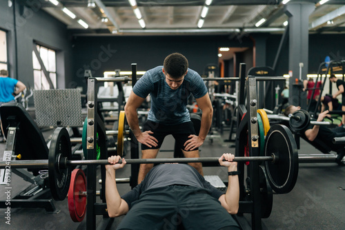 Obraz na plátně Front view of experienced fitness instructor helping beginner sportsman doing barbell bench press exercise during personal workout in gym