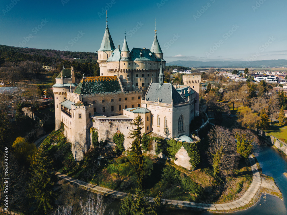 Aerial drone view of Bojnice Castle in Slovakia. High panoramic scenery of medieval Bojnicky Zamok under blue sky, stone walls of fortress with green plants and pine trees, moat with water and hill Aerial drone view of Bojnice Castle in Slovakia. High panoramic scenery of medieval Bojnicky Zamok under blue sky, stone walls of fortress with green plants and pine trees, moat with water and hill