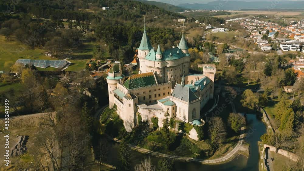 Aerial drone view of Bojnice Castle in Slovakia. High panoramic scenery of medieval Bojnicky Zamok under blue sky, stone walls of fortress with green plants and pine trees, moat with water and hill Aerial drone view of Bojnice Castle in Slovakia. High panoramic scenery of medieval Bojnicky Zamok under blue sky, stone walls of fortress with green plants and pine trees, moat with water and hill
