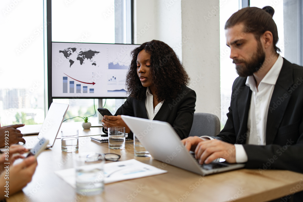 Diverse business team engaged in serious discussion using electronic devices during meeting room presentation, digital charts on screen. Diverse business team engaged in serious discussion using electronic devices during meeting room presentation, digital charts on screen.
