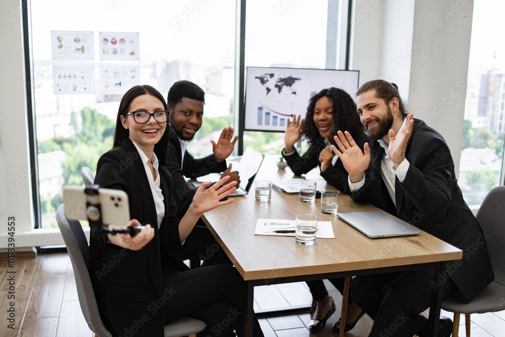Fototapeta premium Smiling diverse business team taking selfie during meeting in modern office. Team members waving at camera while seated around conference table. Professional environment with charts and graphs.