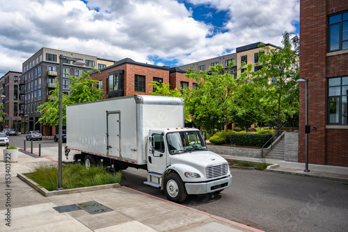 Day cab middle duty rig semi truck with box trailer making delivery standing on the urban street with multistory apartment building