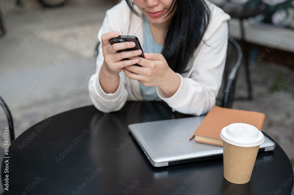 A cropped image of an Asian woman using her smartphone while sitting at an outdoor table of a cafe.