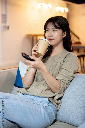 A relaxed Asian woman is having morning coffee while watching TV on the couch in the living room.