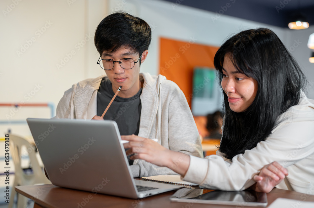 Serious Asian male student is looking at a laptop screen and focusing on listening his friend's idea Serious Asian male student is looking at a laptop screen and focusing on listening his friend's idea