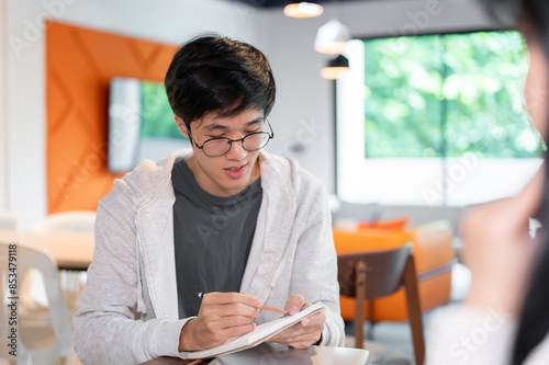 A smart, young Asian male college student is helping his friend with math homework in a coffee shop. © bongkarn