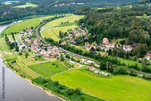 A small town with a river running through it. The houses are small and spread out. The grass is green and the sky is cloudy