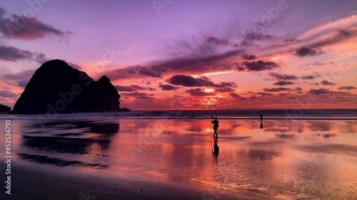 A silhouetted man takes a photo with his camera and tripod on a beach reflecting a beautiful, multicolored sunset on Piha Beach in New Zealand photo