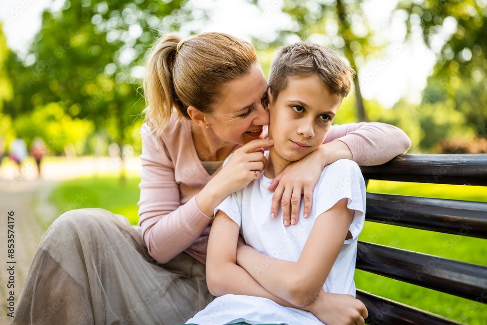 Happy mother is sitting with her son on bench in park. Boy is offended and pouting. Happy mother is sitting with her son on bench in park. Boy is offended and pouting.