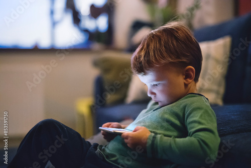 Cute boy sitting on bed watching something on smartphone. Children's screen time. photo