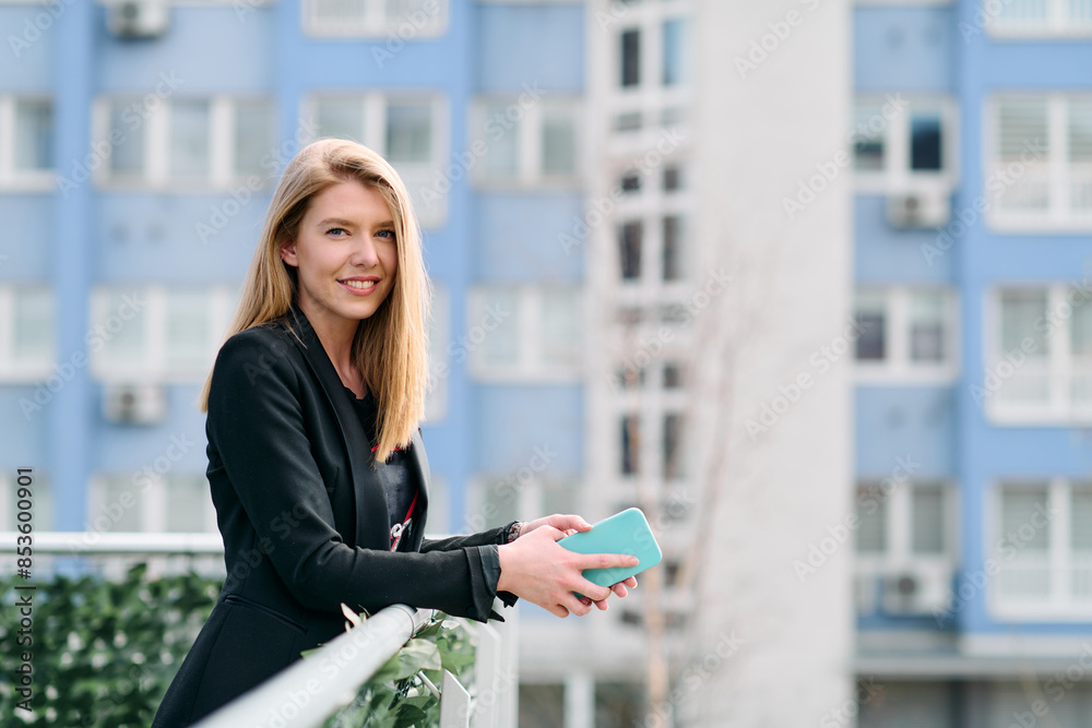 Fototapeta premium Blonde Businesswoman in Black Suit Using Smartphone on Break in Modern Urban Environment.