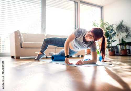 Beautiful redhead woman's home fitness routine with exercise roller. Home workout on gym mt in living room, sporty woman wearing sportswear.