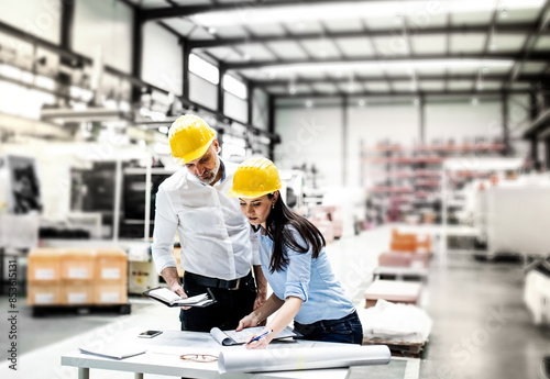 Two project managers standing in modern industrial factory, looking blueprints and documents on table. Manufacturing facility with robotics, robotic arms and automation. Storing products and materials