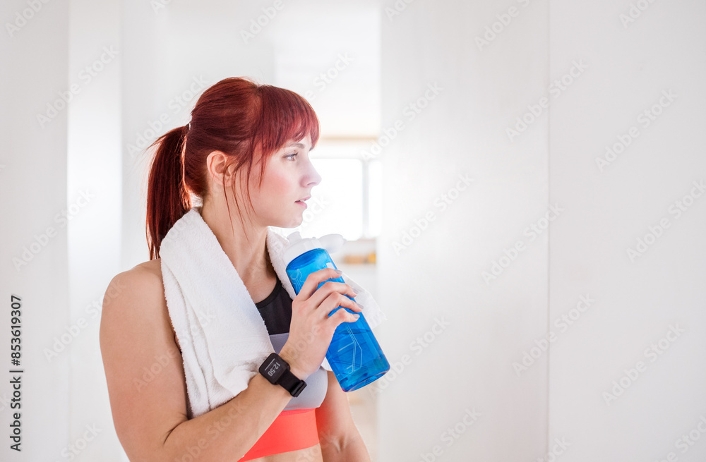 Beautiful redhead woman drinking water, resting after home fitness routine. Home workout on gym mt in living room, sporty woman wearing sportswear. Beautiful redhead woman drinking water, resting after home fitness routine. Home workout on gym mt in living room, sporty woman wearing sportswear.