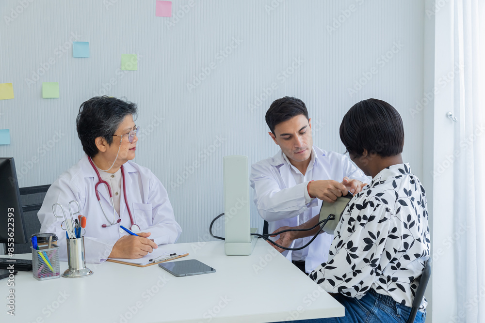 Young Caucasian doctor taking blood pressure of African American woman in clinic. Asian senior female specialist observation. middle-aged patient waiting for diagnosis of disease, medical health care Young Caucasian doctor taking blood pressure of African American woman in clinic. Asian senior female specialist observation. middle-aged patient waiting for diagnosis of disease, medical health care