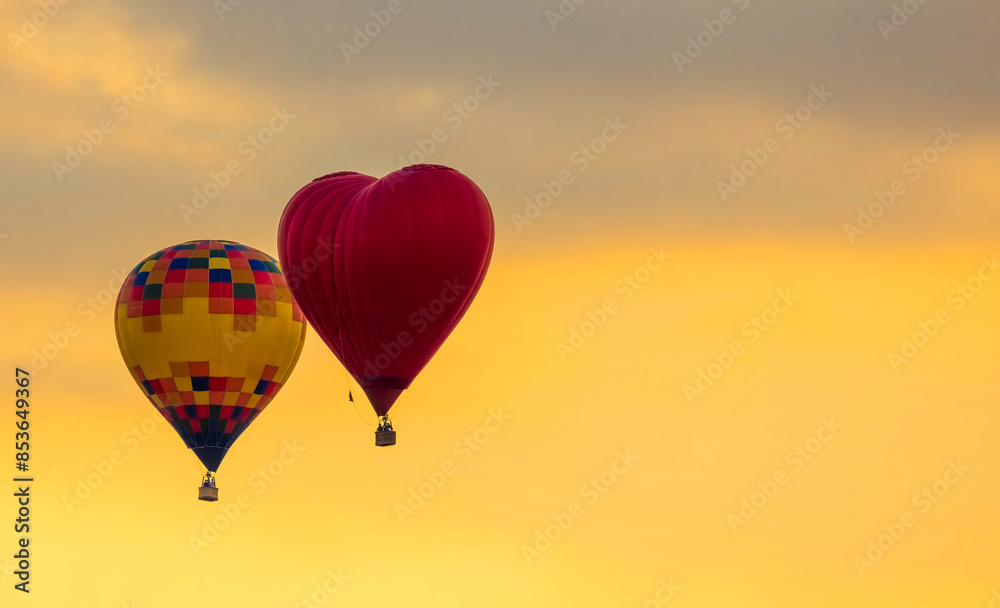 Fototapeta premium Two hot air balloons flying at sunset. the balloon on the right is red and shaped like a heart. The sky is a warm yellow and orange color.