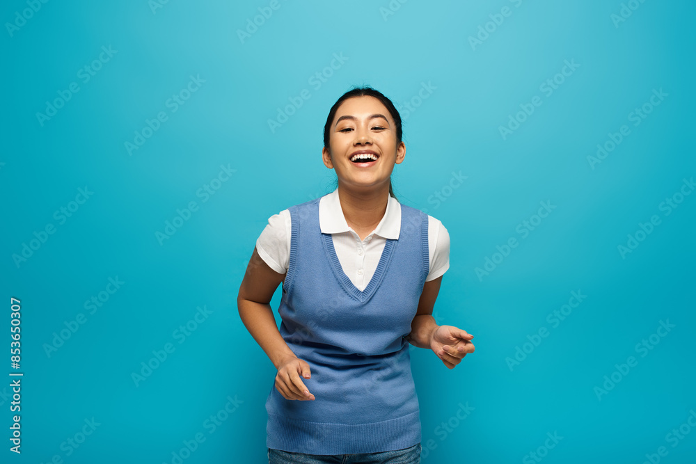 A young Asian woman wearing a white shirt and blue vest smiles brightly against a blue backdrop. A young Asian woman wearing a white shirt and blue vest smiles brightly against a blue backdrop.