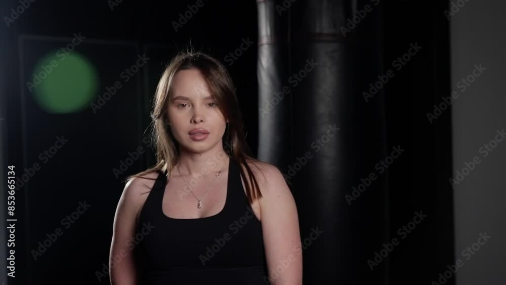 Young woman in black sportswear and boxing gloves, standing in a gym with a punching bag in the background, looking at the camera with confidence and readiness. Young woman in black sportswear and boxing gloves, standing in a gym with a punching bag in the background, looking at the camera with confidence and readiness.