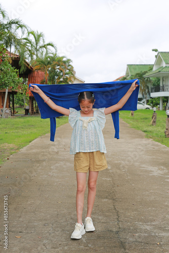 Asian girl child in casual holding a blue sweater over head while standing outdoor at the garden.