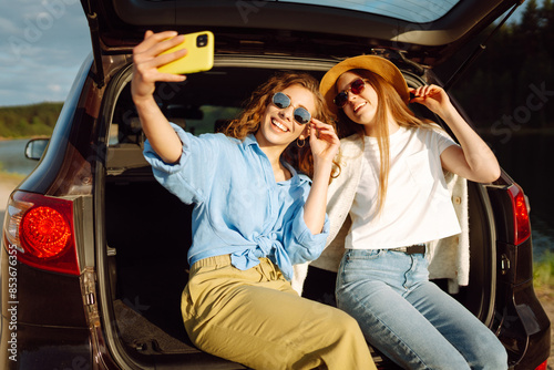Young woman sits in the trunk of a car and takes selfie. Stylish models on a car trip. The concept of technology, blogging, travel.