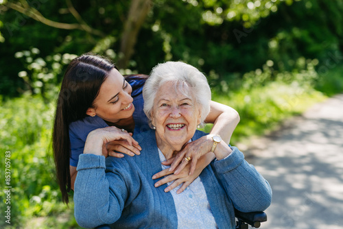 Female caregiver pushing senior woman in wheelchair. Nurse and elderly woman enjoying a warm day in nursing home, public park.