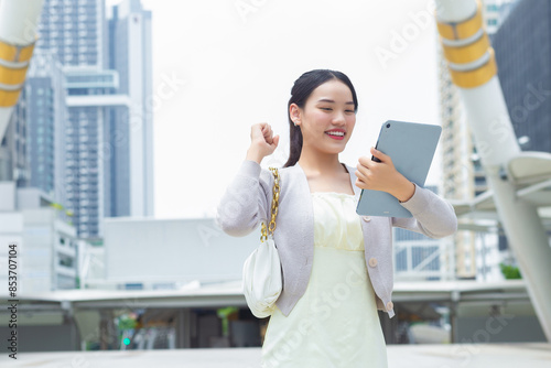 Young Asian business woman is going to office or workplace which she successful while holds notepad in her hands in business while office building in the middle of big city background.
