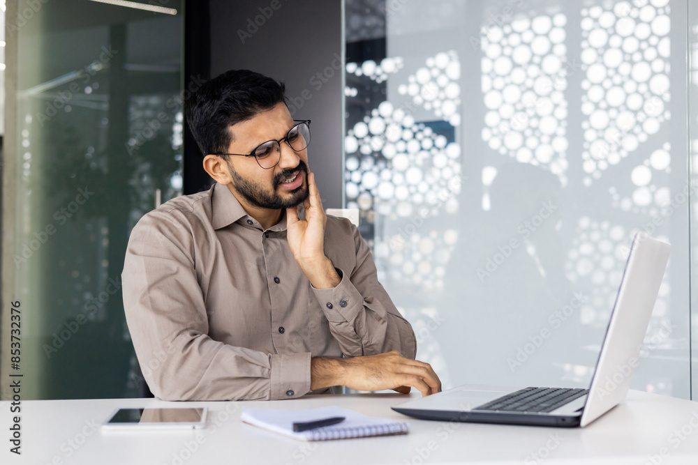 A man suffering from toothache while using a laptop in a contemporary office setting, with pain and discomfort visible on his face. A man suffering from toothache while using a laptop in a contemporary office setting, with pain and discomfort visible on his face.