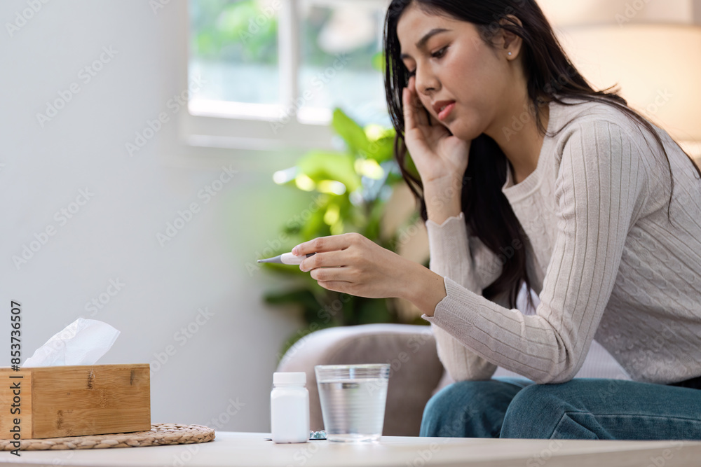 Woman Checking Thermometer While Experiencing Fever and Headache at Home, Sitting on Sofa with Medicine and Tissue Box Woman Checking Thermometer While Experiencing Fever and Headache at Home, Sitting on Sofa with Medicine and Tissue Box