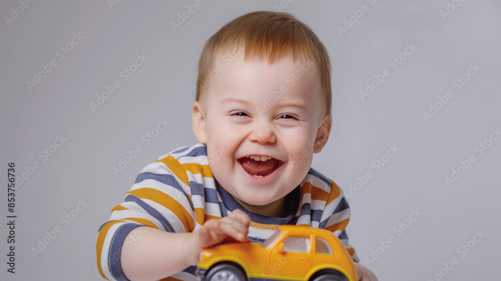 Smiling baby with Down syndrome playing with a yellow toy car while wearing a striped shirt. Joyful expression and playful mood captured on gray background Smiling baby with Down syndrome playing with a yellow toy car while wearing a striped shirt. Joyful expression and playful mood captured on gray background